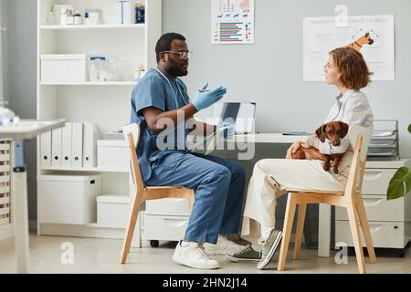 Junge selbstbewusste Tierarzt, der medizinischen Rat an weibliche Tierbesitzer hält niedlichen reinrassigen Dackel auf den Händen Stockfoto