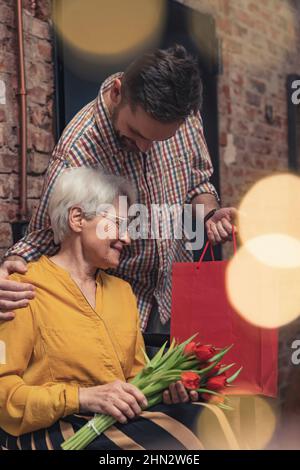 Muttertag Foto eines kaukasischen Sohnes mittleren Alters, der seine grauhaarige Mutter mit einem Geschenk und einem Strauß überraschte. Hochwertige Fotos Stockfoto