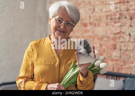Europäische grauhaarige Oma trägt eine Brille und zeigt ein vertikales Foto ihres jüngeren, schönen selbst. Hochwertige Fotos Stockfoto