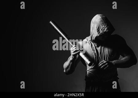 Gefährlicher Mann in der Maske mit Baseballschläger bereit zum Kampf. Ein aggressiver muskulöser Kapuzenmann hält einen Baseballschläger. Stockfoto