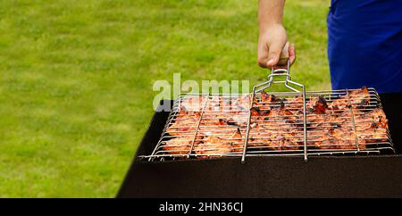 Ein Mann brät auf einem grünen Rasen Kebabs. Ein Mann hält einen Grill mit gegrilltem Fleisch in der Natur. Der Prozess des Kochens von Shish Kebab. Stockfoto