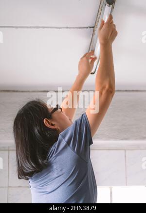 Die Rückseite einer asiatischen Frau mittleren Alters, die die Deckenfluoreszenzlampe selbst wechselt. Das Konzept für Frauen mit Männeraufgaben. Low-Angle-Ansicht. Stockfoto