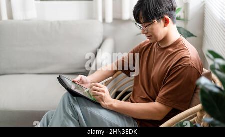 Technologiekonzept der junge Mann, der auf dem Stuhl sitzt und sein iPad hält und etwas darauf notiert. Stockfoto