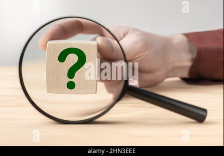 Hand hält Fragezeichen vor der Lupe. Suchen Sie nach einer Antwort auf die Frage. Lupe mit Fragezeichen im Fokus auf Holzwürfel Stockfoto