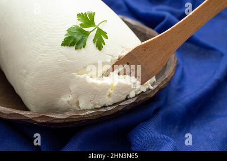 Rumänischer Hüttenkäse auf dem Land, in einer geschnitzten Holzschale mit einer Holzgabel und Petersilienblättern, auf blauer Stoffstruktur Stockfoto