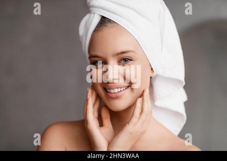 Fröhliche Dame posiert nach dem Baden mit einem Handtuch auf dem Kopf, steht im modernen Badezimmer und lächelt der Kamera zu Stockfoto