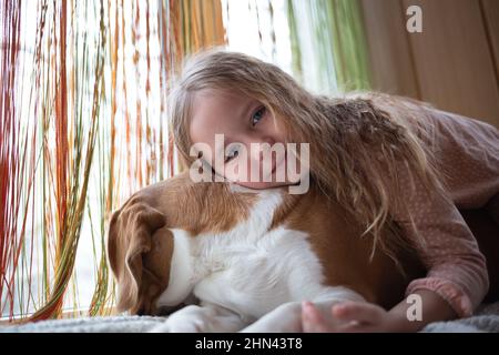 Kleines niedliches Mädchen umarmt einen Beagle-Hund, der auf dem Fensterbrett sitzt Stockfoto