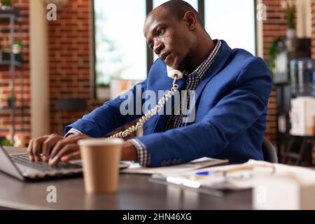 Afroamerikanischer Mann, der Festnetztelefon verwendet, um über die Geschäftsstrategie zu sprechen. Unternehmer im Telefongespräch mit dem Manager, um ein Projekt für einen erfolgreichen Service im Büro zu planen. Stockfoto