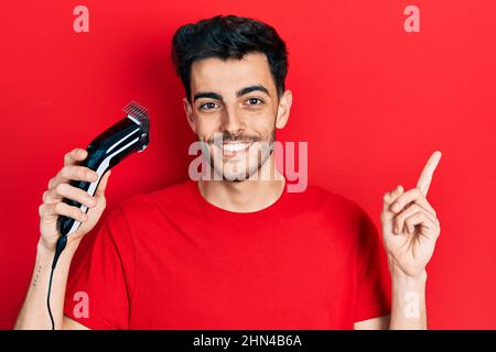 Junger hispanischer Mann lädt elektrische Rasiermaschine lächelnd glücklich zeigt mit Hand und Finger zur Seite Stockfoto