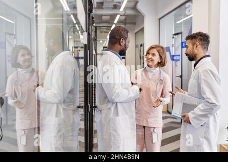 Drei moderne professionelle medizinische Arbeiter in Laborkittel, die bei einem Arbeitstreffen im Korridor der Kliniken diskutieren Stockfoto