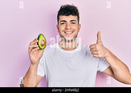 Junger hispanischer Mann mit mittlerer Avocado lächelt glücklich und positiv, Daumen nach oben tun ausgezeichnet und Zustimmung Zeichen Stockfoto