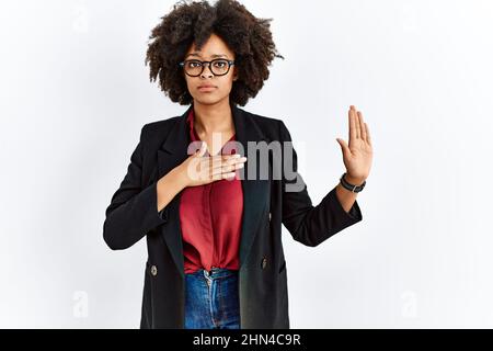afroamerikanische Frau mit Afro-Haaren, die eine Business-Jacke und eine Brille trägt, die mit der Hand auf der Brust und der offenen Handfläche schwört und einen Treueversprechen-Eid ablegt Stockfoto
