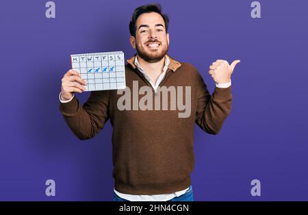 Junger hispanischer Mann hält den Reisekalender und zeigt den Daumen zur Seite und lächelt glücklich mit offenem Mund Stockfoto