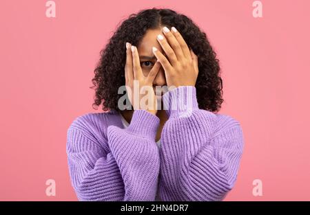 Schockierte junge schwarze Frau, die sich verängstigt fühlt und das Gesicht vor rosafarbenem Studiohintergrund mit Angst bedeckt Stockfoto