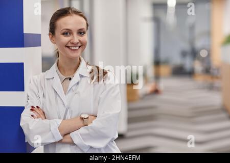 Fröhliche junge Ärztin, die im Labormantel die Arme an der Brust kreuzt, während sie im Korridor moderner Kliniken steht Stockfoto