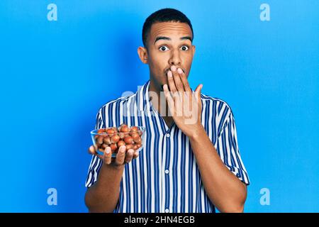 Junger afroamerikanischer Mann mit rohen Haselnüssen, die den Mund mit der Hand bedecken, schockiert und vor Fehlern fürchtend. Überraschter Ausdruck Stockfoto