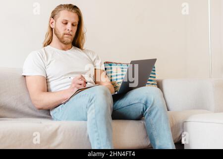 Bärtiger blonder Mann, der mit einem Laptop arbeitet, während er zu Hause auf der Couch sitzt Stockfoto
