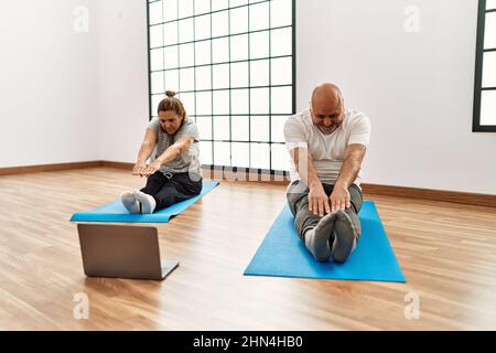 hispanische Paare im mittleren Alter, die sich mit einem Laptop im Fitnessstudio im Online-Sportunterricht ausstrecken Stockfoto