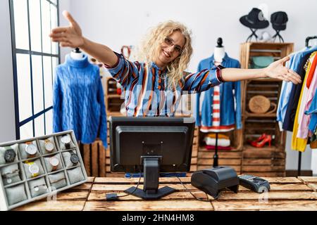 Eine blonde Frau mittleren Alters, die als Managerin in einer Einzelhandelsboutique arbeitet und die Kamera lächelt, um sie mit offenen Armen zu umarmen. Fröhlicher Ausdruck, der Happ umarmt Stockfoto