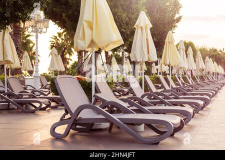 Leere Plastikliegen in einer Reihe und geschlossene Sonnenschirme in der Nähe des Pools bei Sonnenaufgang Stockfoto