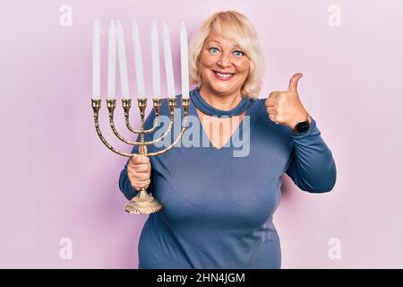 Blonde Frau mittleren Alters, die die Menorah hanukkah jüdische Kerze hält, lächelt glücklich und positiv, Daumen hoch und tut ein ausgezeichnetes und positives Zeichen Stockfoto