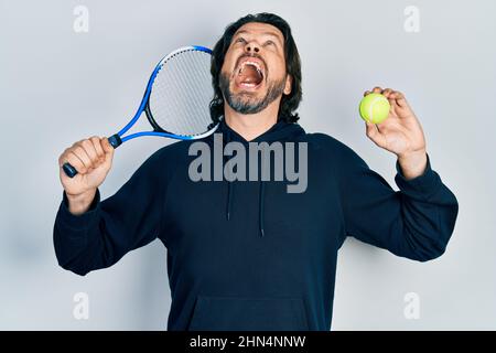 Kaukasischer Mann mittleren Alters spielt Tennis mit Schläger und Ball, wütend und wütend schreiend frustriert und wütend, schreiend mit Wut, die aufschaut. Stockfoto