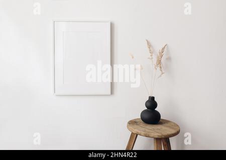 Portrait leerer Bilderrahmen-Mockup in Sonnenlicht. Trockenes festuca, Hasenschwanzgras in moderner schwarzer Bio-Vase. Holzhocker, Stuhl. Weiße Wand Stockfoto