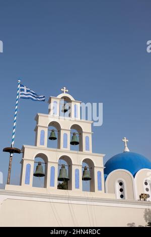 Erstaunliche weiße und blaue griechisch-orthodoxe Kirche mit religiösen Kreuzen, Glocken und der griechischen Flagge, die in der Luft gegen einen blauen Himmel auf Santorini schwenkt Stockfoto