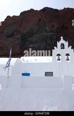 Blick auf eine weiße griechisch-orthodoxe Kirche mit griechischer Flagge und die vulkanische Landschaft des berühmten roten Strandes im Hintergrund Stockfoto