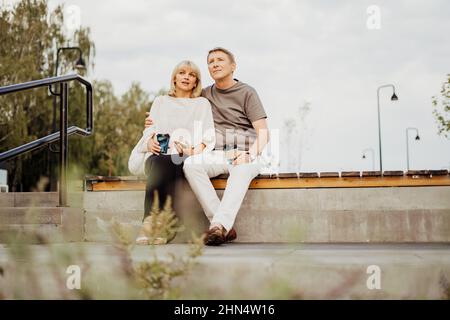 Ein erwachsenes glückliches Paar in der Liebe sitzt auf der Bank im Freien im Stadtpark. Ein blonder Kaukasusmann und eine blonde Frau verbringen Zeit miteinander und trinken Stockfoto