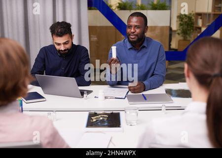Junger, selbstbewusster afroamerikanischer Pharmakologe, der seinen Kollegen eine Plastikflasche mit Pillen zeigt, während er ein neues Medikament vorstellt Stockfoto
