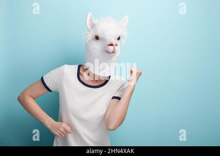 Junge Frau in Alpaka-Maske führen fröhliche unbeschwerte Tanzbewegungen auf blauem Hintergrund aus. Stockfoto