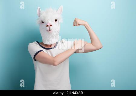 Junge Frau in Alpaka-Maske biegende Armmuskeln, isoliert auf blauem Hintergrund mit Kopierraum. Stockfoto