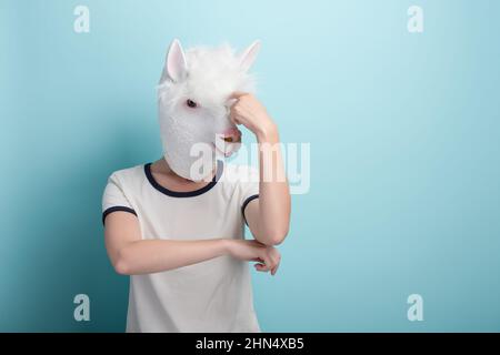 Junge Frau in Alpaka-Maske kratzt sich am Kopf, denkend oder im Zweifel, isoliert auf blauem Hintergrund mit Kopierraum. Stockfoto