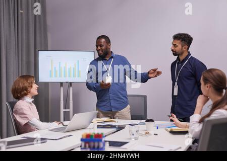 Glücklicher afroamerikanischer Manager oder Experte, der Kollegen neue Mitarbeiter vorstellt, während er sich gegen ein interaktives Whiteboard stellt Stockfoto