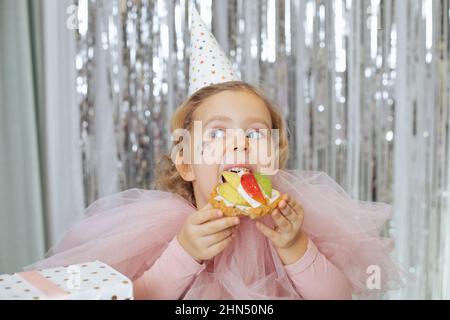 Hübsches kleines Mädchen mit kurzen schönen lockigen Haaren und Sternen im Gesicht in rosa poofy Kleid und Geburtstagskappe essen Obstkuchen. Stockfoto