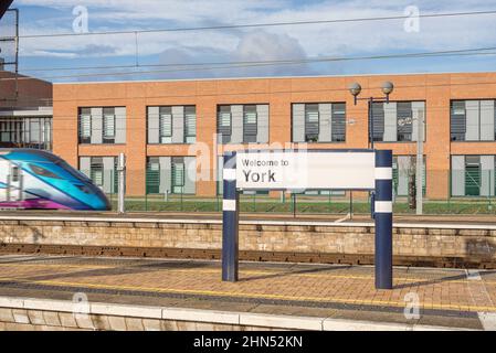 Ein Begrüßungsschild mit Ortsnamen auf einem Bahnhofssteig. Ein modernes Gebäude befindet sich im Hintergrund und ein Zug fährt in den Bahnhof. Stockfoto