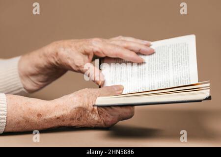 Faltige Hände einer älteren Frau halten ein Buch. Beigefarbener Hintergrund. Das Konzept von Bildung und Lesen Stockfoto