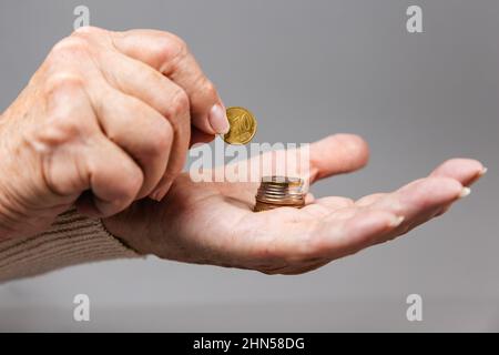Nahaufnahme von Frauen mit faltigen Händen, die eine Euro-Münze in einen Münzstapel gelegt haben. Grauer Hintergrund. Das Konzept der Armut und der kleinen Rente. Stockfoto