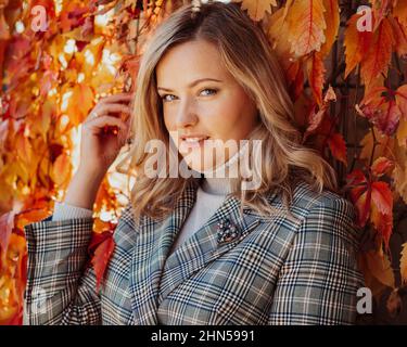 Frau mittleren Alters mit langen welligen blonden Haaren in karierter Jacke und grauem Rollkragen-Pullover posieren berührende Efeu-Blätter. Stockfoto