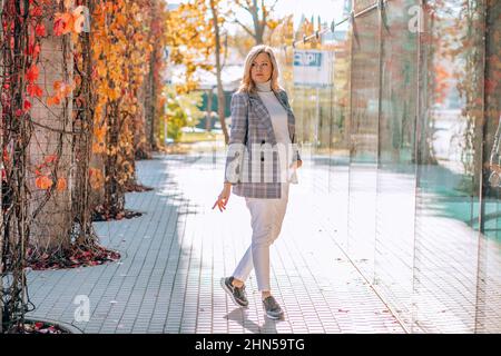 Porträt einer gut aussehenden Frau mittleren Alters in karierter Jacke, Rollkragen-Pullover und weißen Jeans posieren auf Pflasterflagge. Stockfoto