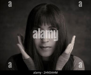 Weibliche Einzelporträts: Attraktive Frau, die direkt auf die Kamera schaut, Hände um das Gesicht. Moody, geheimnisvolles Porträt einer hübschen Dame mit langen dunklen Haaren. Stockfoto