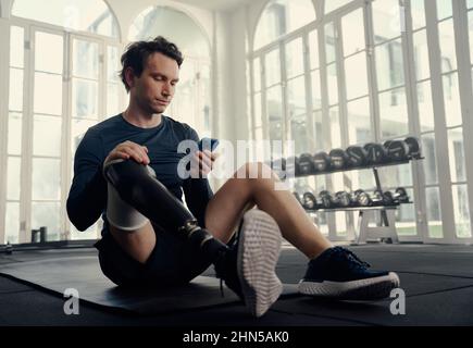 Paralympics-Athlet auf seinem Handy im Fitnesstraining. Mann mit einer Beinprothese auf seinem Handy in einem modernen Fitnessstudio Stockfoto