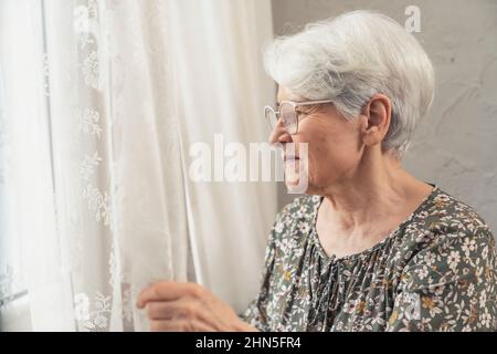 Besorgte grauhaarige kaukasische Großmutter, die mit weißem Vorhang aus dem Fenster schaute. Hochwertige Fotos Stockfoto
