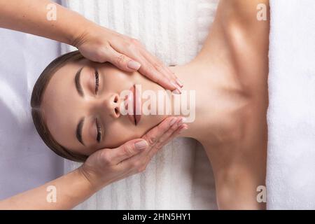 Stress abbauen. Draufsicht auf die schöne junge Frau, die auf dem Rücken liegt, während der Masseur ihr Gesicht massiert Stockfoto
