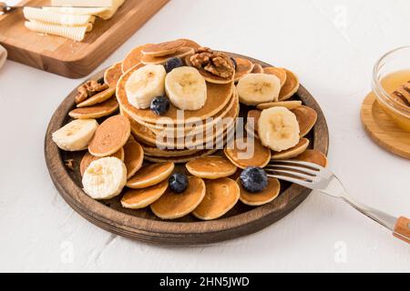Ein großer Holzteller mit Mini-Pfannkuchen und einem Stapel dicker Pfannkuchen in der Mitte. Bananenscheiben, frische Beeren, Nüsse darauf. Weißer Hintergrund Stockfoto