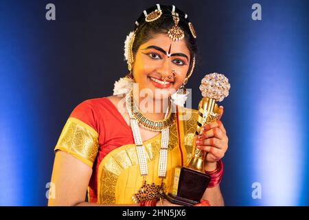 Glücklich lächelnde bharatanatyam-Tänzerin zeigt Siegertrophäe mit Blick auf die Kamera - Konzept der erfolgreichen, erfolgreichen, stolzen und indischen Kultur Stockfoto