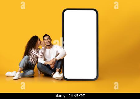 Junges arabisches Paar, das in der Nähe des Smartphones mit weißem Bildschirm sitzt, Frau, die dem Mann von einem coolen Angebot erzählt, sich für das Design vorstellt Stockfoto