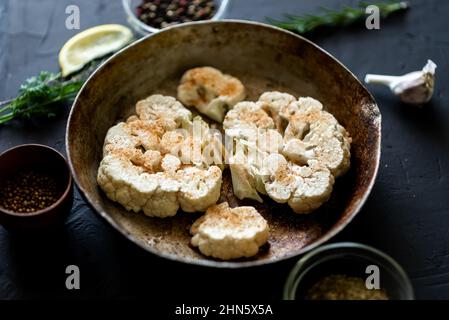 Blumenkohlsteak Kochen. Roher Blumenkohl, mit Gewürzen bestreut, liegt in einer Bratpfanne. Olivenöl, Kräuter, verschiedene Gewürze in der Nähe. Dunkler Hintergrund. Stockfoto