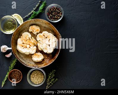 Blumenkohlsteak Kochen. Roher Blumenkohl, mit Gewürzen bestreut, liegt in einer Bratpfanne. Olivenöl, Kräuter, verschiedene Gewürze in der Nähe. Dunkler Hintergrund. Kopys Stockfoto